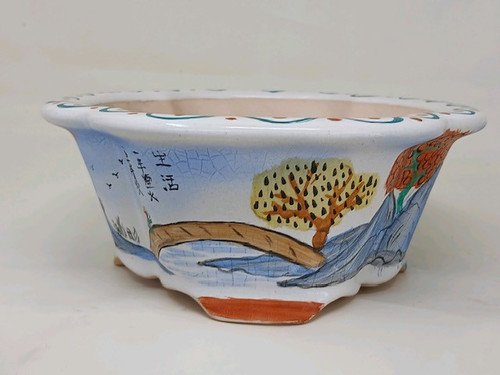 A side-view photo of a 10-inch wide, shallow Yixing bonsai pot. The pot is made of reddish-brown clay, visible at its unglazed base and small feet, while the rest is covered in a white glaze with a delicate crackle pattern. 

The pot features an elegant scalloped, or lotus-style, rim with blue and brown dotted patterns along the edge. Its exterior is adorned with a hand-painted landscape in a traditional Chinese style. On the far left, a column of black calligraphy characters and a few small birds flying in the distance are depicted against the pale blue-tinted background. Centered is a simple arched bridge in brown, spanning a light blue body of water with stylized wave lines. To the right of the bridge stands a vibrant yellow tree with dark spots on its foliage, set against the backdrop of soft blue and grey mountains. The very edge on the right shows the beginning of another tree with dense orange-red leaves. 

The overall aesthetic of this hand-painted bonsai pot is serene and artistic, making it a beautiful and unique container for a specimen bonsai tree.