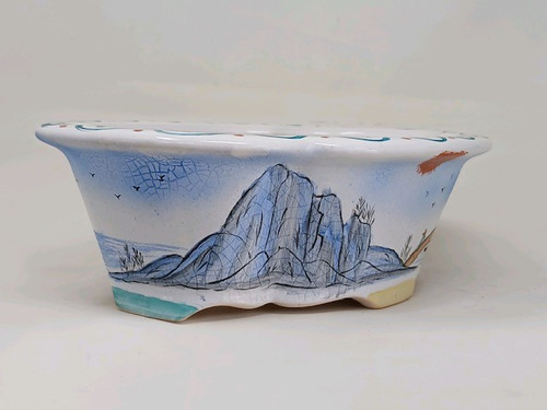 A 10-inch painted Yixing bonsai pot is shown against a plain white background. The pot has a wide, oval shape with a slightly flared rim and is supported by small, scalloped feet. Its white ceramic exterior is adorned with a detailed, hand-painted landscape in a traditional East Asian style. 

Large, blue-tinted craggy mountains with intricate black line work serve as the central motif. To the left, a tranquil water scene with small reeds and a few birds in flight is depicted under a soft blue sky. On the right, a path leads toward a small structure, with more birds and a reddish-brown sun or cloud detail above. 

The entire exterior has a fine crackle glaze, adding a subtle texture and vintage feel. The top rim features a delicate decorative border of blue wavy lines and small reddish dots. The feet of the pot are painted in soft teal and cream colors, providing a colorful base for the elegant landscape artwork. This beautifully crafted Yixing pot is designed to be a functional yet artistic vessel for a prized bonsai tree.