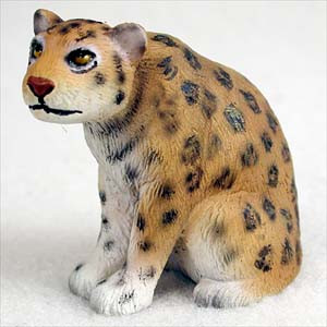 A detailed side-view photograph of a miniature jaguar figurine, specifically designed as an accessory for bonsai trees. The small animal statue is captured in a seated, slightly hunched pose, looking alertly toward the left. Its body is a warm tan or ochre color that fades into a clean white on the chest and paws. The jaguar’s coat is covered in characteristic dark black spots and rosettes, with molded textures throughout the resin to simulate the appearance of fur. Fine details include small rounded ears, bright yellow eyes with black pupils, a pinkish-red nose, and a subtle black line for the mouth. This decorative jaguar figurine is intended to be placed within the landscape of a bonsai tree to add a sense of scale and realism to the miniature garden.