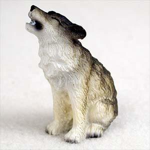 A detailed miniature timber wolf figurine designed as a decorative accessory for a bonsai tree or terrarium. The small wolf is sculpted in a sitting position on its haunches with its head tilted back and mouth open in a classic howl. The figurine features realistic hand-painted fur textures, with a creamy white color on its underbelly, neck, and paws that transitions into dark grey and earthy brown tones along its back, ears, and snout. This wildlife-themed miniature is shown against a neutral, light-grey background, highlighting the intricate details of the timber wolf's pose and features.