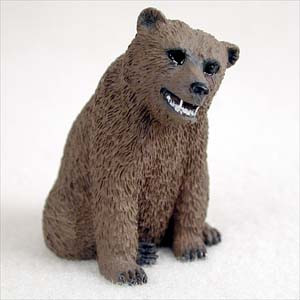 A detailed, close-up photograph of a miniature grizzly bear figurine, designed as a decorative accessory for a bonsai tree or fairy garden. The brown bear is depicted in a seated position, leaning slightly forward and to its right. Its entire body is covered in a realistically molded texture that mimics thick, coarse fur. The bear's facial expression features small, dark, bead-like eyes and a black nose, with its mouth slightly open to reveal a row of white teeth in a subtle snarl or grin. Its front and back paws are visible at the base, featuring dark, pointed claws. The figurine is set against a clean, off-white neutral background, emphasizing the fine craftsmanship of the small animal statue.