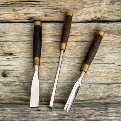 A top-down view of three Tinyroots premium wood carving tools laid out on a rustic, weathered wood background. This heavy-duty 3-piece set includes a wide flat chisel, a narrow square-tipped chisel, and a curved gouge, perfect for intricate bonsai work like deadwood carving on large trees or Yamadori. Each tool features a gleaming, polished steel blade and a dark, cylindrical wooden handle. The handles are elegantly finished with brass ferrules at both ends, emphasizing the high-quality craftsmanship of these professional bonsai tools.