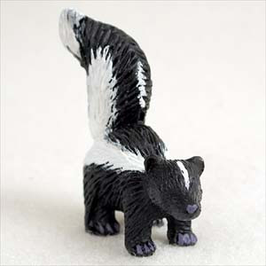 A miniature skunk bonsai tree figurine stands on a neutral, off-white surface in a close-up product shot. This small animal figurine features a classic black and white color scheme, with the black portions of its body intricately textured to resemble thick fur. A broad, bright white stripe begins between its tiny rounded ears, runs down its back, and covers the underside of its large, arched, bushy tail, which is held high in the air. 

The skunk is depicted on all fours in a playful stance, looking slightly to the side. Notable artistic details include a small lavender-purple nose and matching purple-colored paws that contrast with its monochrome body. Designed as a whimsical garden ornament, this miniature skunk is scaled perfectly to add a touch of personality and nature-inspired charm to an indoor bonsai tree arrangement or a miniature fairy garden.