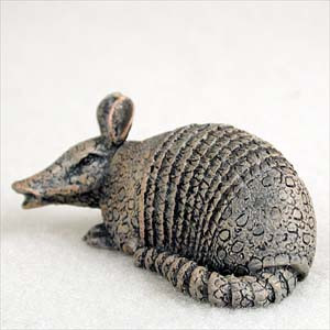 An intricate miniature armadillo bonsai tree figurine, shown in a side profile against a plain, light-colored background. This detailed sculpture is designed as a bonsai pot accessory and features a realistic, textured finish that resembles aged bronze or stone. The armadillo is depicted in a low, crouching pose with its long snout slightly open, prominent upright ears, and a thick, segmented tail curled along its side. Its protective armor is rendered with fine detail, showing characteristic horizontal bands across its back and a pebbled skin texture on its head and sides, making it a charming and characterful addition to a miniature landscape or bonsai arrangement.
