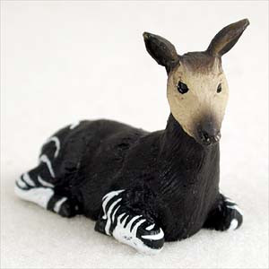A detailed miniature okapi figurine designed as an accessory for a bonsai tree arrangement. The small animal is depicted in a resting, lying-down position. It features a dark brown body with the species' signature black and white horizontal stripes on its upper legs and haunches. The figurine has a light tan face, small black eyes, and large, upright ears, with a textured surface that gives it a realistic appearance. The piece is photographed against a clean, white background, emphasizing its fine craftsmanship.