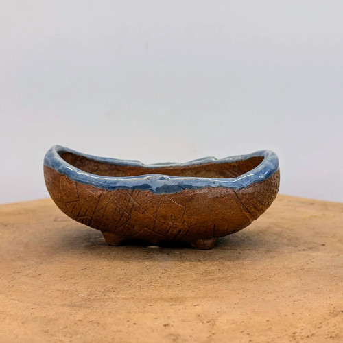A side-profile view of a small, oblong handmade bonsai pot created by American ceramic artist Bruce Lenore. The pot is crafted from a warm, reddish-brown clay with a deeply textured exterior featuring geometric, hand-etched lines and cross-hatching that give it a rustic, organic feel. 

The top edge of the pot has an undulating, wavy rim finished in a thick, glossy sky-blue glaze that pools and drips slightly down the textured sides. The piece is boat-shaped and stands on small, integrated ceramic feet, elevating it above the flat, light-tan wooden surface it sits upon. The background is a clean, minimalist white, highlighting the artisanal craftsmanship and unique color contrast between the earthy brown clay and the vibrant blue glazed rim.