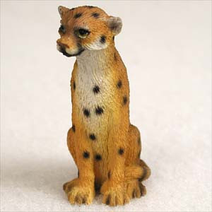 A detailed miniature figurine of a cheetah, designed as an accessory for a bonsai tree or miniature landscape. The small statue depicts the cheetah sitting alertly on its haunches, looking slightly to its left with a focused expression. The figurine is crafted with a textured surface to mimic the appearance of fur and is hand-painted in a warm golden-orange hue with characteristic black spots scattered across its body. A lighter, cream-colored patch runs down its chest and underbelly. The cheetah's face features small, rounded ears, dark, lifelike eyes, and a black nose, adding to its realistic charm. It is set against a plain, neutral-toned background that emphasizes its detail and small scale, making it a perfect decorative element to add a touch of the wild to a bonsai arrangement.