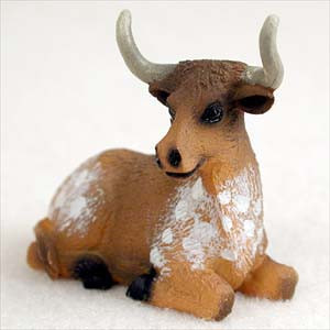 A close-up shot of a miniature longhorn steer figurine, designed as a decorative accessory for bonsai tree arrangements. The small animal figure is shown in a peaceful resting position, lying down with its legs tucked underneath and its head turned slightly to the side. The steer's body is a warm, tawny reddish-brown color, accented with hand-painted white spots and patches across its shoulders and back. It features two prominent, upward-curving horns in a light greyish-white hue, dark black hooves, and glossy black eyes that give it a life-like expression. The figurine is set against a soft-focus, textured white background, highlighting its intricate detail and small scale.