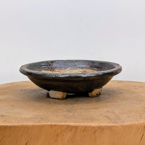 A handcrafted ceramic bonsai pot by American artist Bruce Lenore, showcased on a light wood display stand. This shallow, round pot features a rustic wabi-sabi aesthetic with a dark, textured metallic glaze that contrasts against areas of exposed, light-colored sandy clay on the interior and base. The pot is supported by three small, blocky feet that reveal the raw clay's natural, earthy tones. Its low profile and artisanal finish make it a unique and professional container for bonsai enthusiasts. The image is set against a simple off-white background, highlighting the intricate details, varying glazes, and earthy textures of the handmade piece.