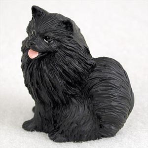 A detailed miniature figurine of a solid black Pomeranian dog, designed as a charming decorative accent for bonsai tree displays or indoor gardens. The figurine captures the dog in a sitting position, facing slightly to the left with a cheerful expression. It features intricate, deeply carved textures that realistically mimic the Pomeranian's characteristic thick, fluffy coat and long fur. The dog has small, alert, pointed ears, dark shining eyes, and a tiny pink tongue sticking out of its slightly open mouth. The entire piece is rendered in a matte black finish and is photographed against a neutral, light-colored background to highlight its silhouette and fine details as a pet-themed accessory.