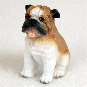 A detailed miniature figurine of an English Bulldog, sitting upright and looking slightly to the left. The realistic figurine features a brown and white coat with a molded, fur-like texture. Its characteristic bulldog face includes a wrinkled brow, black muzzle, dark eyes, and a small pink tongue sticking out in a friendly expression. The head and back are primarily brown, while the face, chest, and legs are white. This charming, small dog statue is designed as an accent piece or accessory for a bonsai tree display or miniature garden.