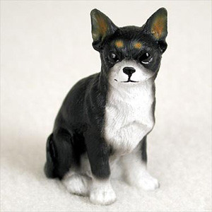 A detailed miniature figurine of a black and white Chihuahua, captured in a sitting, attentive pose and facing directly forward. The figurine features a primarily black coat on its head and back, which is sharply contrasted by a white muzzle, chest, and front paws. The iconic physical traits of the breed are well-defined, including large, upright pointed ears with hints of tan inside, round expressive dark eyes, and small tan dots above each eye resembling eyebrows. The surface of the resin figurine is subtly textured to mimic the look of real fur. Set against a neutral, off-white, softly textured background, this small Chihuahua figurine is designed as a decorative accessory for a bonsai tree arrangement or a miniature garden display.