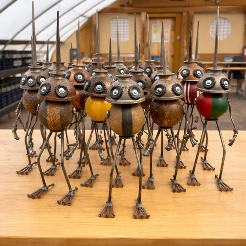 A large group of whimsical, handcrafted found-object robot sculptures stands assembled on a light-colored wooden tabletop. Each unique robot is meticulously crafted from vintage materials, featuring a weathered wooden croquet ball for a body and an antique metal oil can as a head. The oil cans are inverted, with their long, thin spouts pointing straight upward like metallic antennae or spikes.

The robots have expressive, wide-set eyes made from hardware like washers and screws, giving them a curious and friendly appearance. Their spindly arms and legs are constructed from thin metal rods, ending in small, multi-pronged feet and hands made of tiny screws. The croquet balls used for the bodies vary in color and design; some are solid yellow, others feature natural wood grain with painted stripes, and one prominently displays a split red and green pattern.

The sculptures are arranged in a staggered group, as if posing for a portrait. The background reveals a greenhouse-like setting with a translucent arched roof and wooden framing, suggesting an artisan workshop or a display at a bonsai nursery. The warm, natural lighting highlights the rustic textures of the aged wood and the metallic sheen of the repurposed parts.