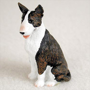 A detailed miniature figurine of a Bull Terrier, meticulously painted with a brindle and white coat. The figurine captures the breed's distinctive egg-shaped head, pointed pricked ears, and small dark eyes. This Bull Terrier is posed in a sitting position, looking slightly to the left with its mouth ajar, showing a hint of a pink tongue. 

The brindle pattern, a mix of dark brown and black streaks, covers the top of its head and most of its back and tail, while its muzzle, chest, and front legs are a crisp white. The surface of the figurine is textured to mimic the look of short, coarse fur. Set against a neutral, off-white background, this small collectible is designed as a bonsai tree accessory, intended to add a lifelike and charming element to a miniature garden or indoor bonsai display.