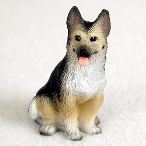 A detailed miniature figurine of a German Shepherd dog, designed as a decorative accessory for a bonsai tree or miniature garden. The figurine depicts the dog in a sitting position, alert and facing forward with its pink tongue playfully poking out. It features realistic coloring characteristic of the breed, with a black saddle over its back, a black mask around its eyes and snout, and a tan-colored face and legs. Touches of white fur are visible on its chest and paws. The dog's tall, pointed ears have dark edges and pinkish interiors, and its small, dark eyes have a slight shine. The figurine has a textured surface to resemble fur and is set against a plain, soft white background.