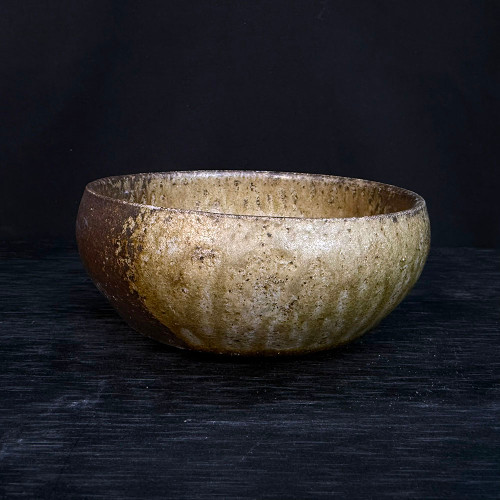 A studio photograph of a 5.5-inch handmade bonsai pot by artist Clayton Lanphear (No. 046). The low-profile, round ceramic pot features a rustic, heavily textured glaze with an earthy palette of mottled beige, sandy tan, and deep umber. Subtle vertical glaze drips and a pitted, organic surface give the pot a weathered, wabi-sabi aesthetic. The vessel is centered on a dark, horizontally-grained wooden surface and set against a solid black background, with soft lighting that emphasizes its hand-crafted form and unique textural details.