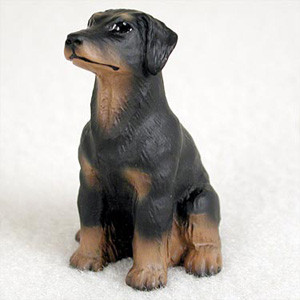 A detailed miniature figurine of a Doberman Pinscher puppy, designed as a decorative companion for a bonsai tree or miniature garden. The dog is depicted in a sitting pose, looking alertly upward and toward the left. It features the classic Doberman color pattern, with a primarily matte black body and tan (rust) markings on its muzzle, chest, and paws. This specific figurine shows the breed with natural, uncropped ears that hang down the sides of its head. The surface of the figurine has a fine, molded texture that mimics the appearance of short canine fur. It is set against a plain, off-white textured background.