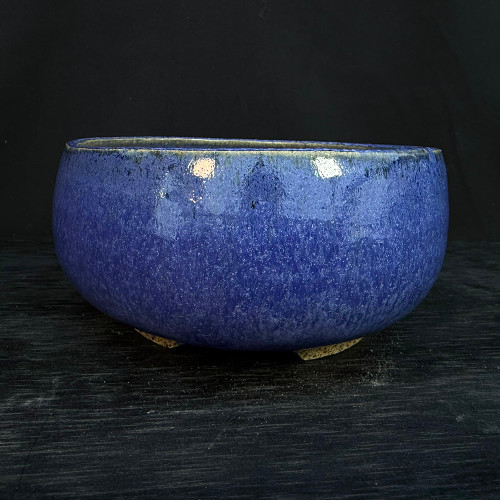 A detailed studio photograph shows a 5.75-inch handmade ceramic bonsai pot, crafted by artist Clayton Lanphear. The pot features a rounded, bulbous shape with a slightly inward-tapering rim, finished in a rich, glossy cobalt blue glaze. The glaze is characterized by a dense, mottled pattern of lighter blue and white speckles, creating a textured visual effect similar to a starry night sky. The pot's dark rim transitions into the vibrant blue body, and it sits elevated on three small, unglazed, light-tan ceramic feet. It is displayed against a dark, textured charcoal-colored surface and a solid black background, which emphasizes the pot's deep color and reflective, high-shine finish.