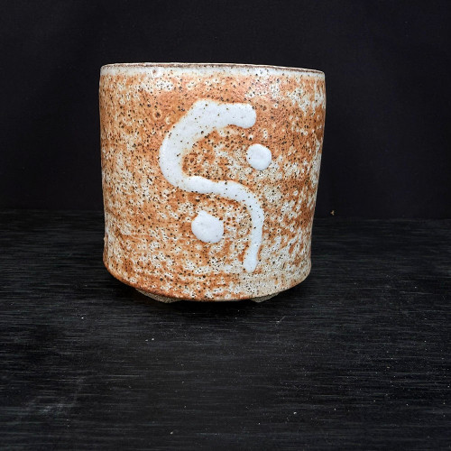 A detailed studio shot of a 3.25-inch handmade ceramic bonsai pot by artist Clayton Lanphear. This wood-fired vessel displays a cylindrical shape with a rustic, heavily textured surface. The exterior features a light cream base densely covered in warm, toasted-orange and dark brown speckling, characteristic of the wood-firing process. A prominent, raised white abstract design is centered on the front, consisting of a thick, curved 'S' shape accented by two solid white circles, reminiscent of a minimalist yin-yang motif. The pot sits on a dark, horizontally grained surface, set against a deep, non-reflective black background that emphasizes the pot’s earthy tones and artisanal craftsmanship.