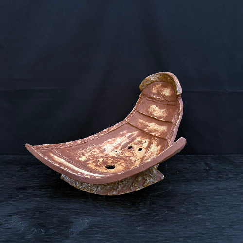 An overhead, slightly angled view of a 15-inch handmade ceramic bonsai planter created by artist John Fazzino. The planter features a unique, organic slab-built design with a sweeping crescent or boat-like shape, where both ends curve upward dramatically. It is crafted from a deep reddish-brown terracotta clay, accented with irregular patches of creamy tan and beige glaze in the center and along the interior ridges, giving it a weathered, rustic appearance. 

The interior of the pot shows visible horizontal slab marks, while the exterior base has a rugged, craggy texture reminiscent of tree bark. Functional elements are visible at the bottom, including a large central drainage hole and several smaller holes intended for anchoring bonsai wire. The planter sits on a dark, textured charcoal-colored surface against a solid black background, which emphasizes the warm, earthy tones and handcrafted texture of the ceramic art piece.