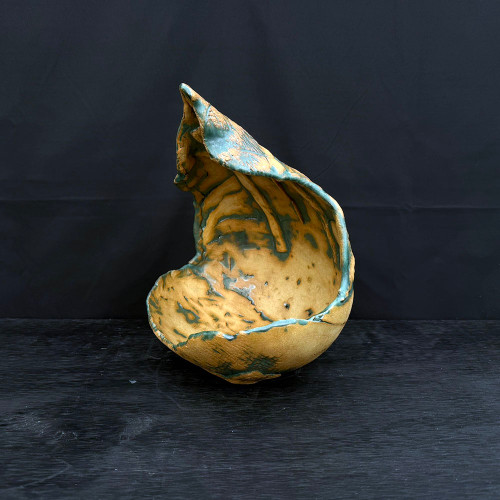 A 13-inch handmade crescent-shaped ceramic bonsai planter by artist John Fazzino, labeled as No. 004. This sculptural piece features a dramatic, organic form that tapers to a point, resembling a piece of naturally weathered bark or a large, curled leaf. The surface is heavily textured with an earthy ochre and sandy yellow base color, accented by a teal or sea-foam green glaze that pools in the ridges and lines the jagged, irregular edges. The planter is photographed against a deep black background, resting on a dark, wood-grained surface, highlighting its unique, rustic silhouette and wabi-sabi aesthetic.