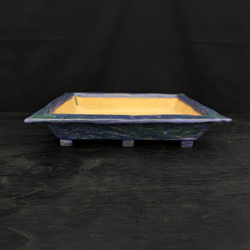 A side-view photo of a handmade, rectangular ceramic bonsai planter by artist John Fazzino. This shallow, artisanal pot features a unique, multi-tonal glaze on its exterior, blending deep shades of indigo blue with vibrant forest green. The surface is heavily textured with organic, bark-like ridges and indentations, giving it a rugged, naturalistic feel. A wide, outward-flaring rim frames the top, revealing a warm, unglazed yellowish-orange clay interior. The pot is supported by several small, square feet and is presented on a dark, weathered wood surface against a solid black background, highlighting its intricate craftsmanship and bold color palette.