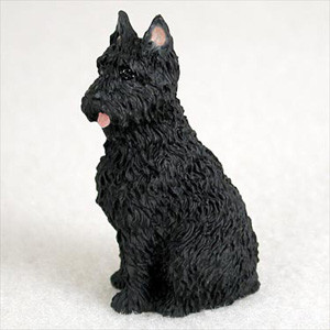 A detailed miniature figurine of a black Bouvier des Flandres dog, designed as a decorative accessory for a bonsai tree. The figurine features a highly textured, shaggy coat characteristic of the breed, with a thick beard and bushy eyebrows. It is depicted in a sitting pose, looking slightly to the side with alert, pointed ears and a small pink tongue peeking out. The figurine is set against a plain, light-colored background, highlighting its intricate sculptural details.