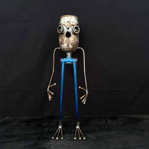 A whimsical handcrafted robot sculpture, part of a limited edition series, stands centered against a solid black background. The piece is fashioned from various reclaimed metal parts, showcasing an industrial-chic and upcycled aesthetic. Its head is a tall, cylindrical metal canister with a heavily rusted and weathered finish, featuring two protruding round metal pieces for eyes and a small circular hole for a mouth, giving the robot a curious expression. Thin, silvery metal arms curve outward and down from the "neck" area, ending in hands made from five fanned-out rusted screws that resemble fingers. The robot's long, spindly legs are made from a tapered blue metal component, possibly a bicycle fork, which shows signs of wear and chipping paint. Its feet are crafted from several metal rods with rounded heads, fanned out like toes to provide a stable base on the dark, textured surface where the sculpture stands.