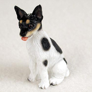 A detailed miniature figurine of a Rat Terrier dog, designed as a charming decorative accent for a bonsai tree display or a fairy garden. The small dog is captured in a sitting position, featuring a primarily white coat with distinct black patches on its shoulder and flank. Its head is predominantly black with characteristic tan "eyebrow" markings and a tan muzzle, while its alert ears stand upright. A small, pink tongue playfully pokes out from its mouth, adding a touch of personality. The figurine's molded surface subtly mimics the texture of short fur, making it a realistic and whimsical addition to any miniature landscape or indoor garden setting.