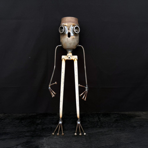 A full-shot, studio photograph of a unique, limited edition robot sculpture (identified as No. 207) crafted from various reclaimed and scrap metal components. 

The sculpture features a tall, slender, and whimsical design. Its head is a rounded, weathered metal canister with an industrial patina, featuring two large, protruding eyes made from metal rings and a small, circular hole for a mouth, giving it a curious expression. The robot's thin arms are made from bent metal rods that end in hands fashioned from several screws welded together to look like fingers.

The lower half of the sculpture consists of long, thin legs made from white-painted metal tubes—resembling parts of a bicycle fork—which connect at the top to form a narrow hip. Each leg ends in a splayed foot made of five metal pins with rounded ball tips, reminiscent of a bird's foot. 

The entire reclaimed metal art piece stands upright on a dark, textured wooden or metallic floor against a solid, matte black background. The lighting emphasizes the various metallic textures, weld marks, and rust-colored accents, highlighting its rustic, industrial aesthetic. This decorative sculpture is designed as a companion piece for bonsai trees or home gardens.