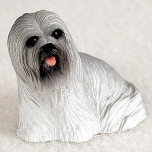 A small, intricately detailed Lhasa Apso dog figurine designed as a charming accessory for a bonsai tree display. The figurine is crafted with a realistic texture to mimic the breed's signature long, flowing coat in shades of gray and white. It features a dark face with shiny black eyes, a black nose, and a playful pink tongue peeking out. The dog is in a sitting or crouching pose, looking off-center towards the viewer's left. The entire piece is set against a neutral, light-colored background, emphasizing its delicate features and suitability for enhancing a miniature landscape or indoor garden.