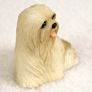 A detailed miniature figurine of a blonde Lhasa Apso dog, designed as a decorative accessory for a bonsai tree arrangement. The small sculpture captures the breed's iconic long, flowing coat with intricate, carved textures in a pale tan or cream color. The dog is depicted in a sitting position, looking off to the side with dark, expressive eyes, a small black nose, and a tiny pink tongue peeking out from its mouth. The figurine is photographed against a soft, neutral, out-of-focus background that emphasizes the fine details of the miniature's craftsmanship.
