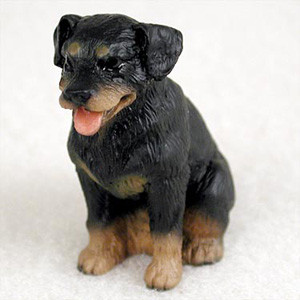 A detailed miniature figurine of a Rottweiler puppy, designed as a decorative accessory for a bonsai tree or terrarium. The figurine captures the breed's classic features, with a sleek black coat and characteristic tan markings above the eyes, on the muzzle, chest, and sturdy paws. It is depicted in a sitting position, looking slightly to the side with an alert and friendly expression. The puppy’s mouth is open in a happy pant, revealing a small, soft pink tongue. The figurine's surface is intricately textured to mimic the appearance of short, dense fur. It is shown centered against a plain, off-white background that highlights the craftsmanship and coloring of the small-scale dog model.
