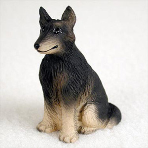 A detailed Belgian Tervuren figurine designed as a bonsai tree accessory. The miniature dog is depicted in a sitting position, looking slightly off to the side with a calm expression. It features the breed's distinctive coloring: a black mask on its muzzle, dark ears, and a black "saddle" of fur over its back, which contrasts with its light tan or beige chest, legs, and underbelly. The surface of the figurine is finely textured to replicate the appearance of long, thick dog fur. Set against a soft, plain white background, this small handcrafted figurine is intended to be placed in the soil of a bonsai pot to add life, personality, and a sense of scale to a miniature tree display.