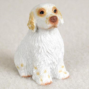 This detailed miniature figurine depicts a Clumber Spaniel dog, designed as a decorative accessory for a bonsai tree, terrarium, or fairy garden. The figurine is captured in a patient, upright sitting position, looking slightly upward and to the side with a gentle and alert expression. 

True to the breed's characteristics, the spaniel is primarily white with light lemon-colored markings. These tan accents are visible on its long, floppy ears, in patches surrounding its dark, rounded eyes, and as small freckles across its muzzle and on its front paws. A prominent, smooth brown nose sits centered on its face. 

The entire surface of the miniature is intricately molded with ridges and grooves to realistically mimic the texture of thick, wavy fur along its chest, back, and feathered legs. Photographed against a soft, neutral-colored background, this small Clumber Spaniel figurine provides a touch of life-like detail for indoor miniature landscapes.