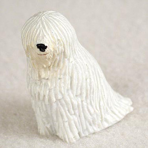 A detailed miniature Komondor bonsai tree figurine, meticulously sculpted to capture the breed's iconic corded white coat. The small, cream-colored dog statue features vertical grooves mimicking thick dreadlocks that cover its body and drape over its face, leaving only a tiny black nose and a faint hint of a mouth visible. This charming animal miniature is shown sitting on a light-colored, textured surface, designed to be used as a decorative accent for a miniature garden, terrarium, or bonsai tree display.