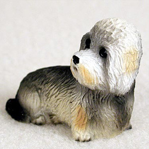 A detailed close-up photograph of a Dandie Dinmont Terrier bonsai tree figurine. The miniature dog statue is captured in a resting position, lying down on its belly with its head turned alertly to the side. The figurine features the breed's distinctive characteristics, including a textured, pepper-grey and tan coat and the iconic large, silky white topknot of hair on its head. Finely painted details highlight its dark, expressive eyes and black nose, giving the small sculpture a lifelike appearance. Designed as a charming accessory for miniature landscaping, this Dandie Dinmont figurine adds a whimsical and personalized touch to a bonsai garden or indoor plant display. The figurine is set against a neutral, off-white textured background.