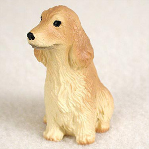 A detailed miniature figurine of a blonde English Cocker Spaniel, designed as a decorative accessory for a bonsai tree. The small dog figurine is depicted sitting upright in a classic pose, looking slightly towards the left. It features the breed's hallmark long, floppy ears with wavy, sculpted fur, a prominent black nose, and dark, soulful eyes. The color of the figurine is a light golden-blonde, with paler, cream-colored highlights on the chest and underbelly, meticulously textured to represent feathered fur. The figurine rests on a plain, textured off-white surface, highlighting its intricate craftsmanship and charm as a pet-themed garden ornament.