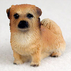 A realistic miniature Tibetan Spaniel figurine designed as a bonsai tree accessory. The small dog statue is rendered in a honey-tan or golden-brown color with highly detailed, textured fur that captures the breed's characteristic thick ruff around the neck and feathered coat. The figurine is depicted in a standing pose, looking forward with dark, soulful eyes, a prominent black nose, and small, floppy ears that are shaded with a darker brown at the tips. Its bushy, plume-like tail is curled over its back, a distinctive trait of the Tibetan Spaniel. The craftsmanship emphasizes a lifelike quality, making it a charming addition to a miniature landscape or the base of a bonsai tree. The figurine is set against a neutral, off-white background that highlights its intricate details and coloring.