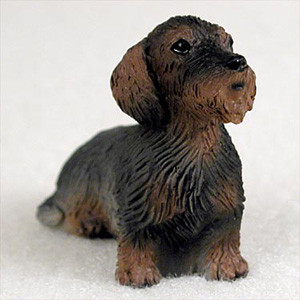 A small, detailed figurine of a wire-haired dachshund designed as an accessory for a bonsai tree. The miniature dog is depicted in a seated pose, facing towards the right with an alert expression. Its coat features a realistic 'wild boar' coloration, primarily dark black along the back with deep reddish-brown accents on its chest, legs, ears, and snout. The surface is intricately sculpted with deep, vertical ridges to recreate the coarse, wiry texture characteristic of the wire-haired breed. It has long, drooping ears and a prominent, textured beard and mustache around its muzzle. The figurine is set against a plain, soft-focus white background, emphasizing its fine craftsmanship.