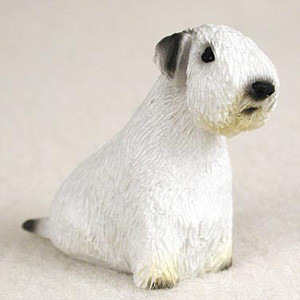 A detailed, hand-painted miniature Sealyham Terrier figurine, specifically designed as a decorative accessory for bonsai trees or miniature gardens. The small dog figurine is predominantly white with a textured finish that captures the appearance of a terrier's wiry coat. Its characteristic breed features include a long, rectangular head, small dark eyes, a prominent black nose, and dark grey ears that fold forward. The figurine is depicted in a seated position, shown from a three-quarter side profile. A small, dark-tipped tail nub is visible at the rear, and subtle tan shading appears near the paws for realistic detail. The figurine rests on a neutral, off-white surface, highlighting its intricate details and suitability as a charming accent piece for indoor or outdoor bonsai displays.