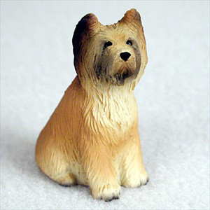 A small, detailed figurine of a Briard dog, designed as a decorative accessory for a bonsai tree or miniature garden. The figurine depicts the dog in a sitting position, looking alertly upwards and to the side. It features a long, shaggy coat, intricately sculpted to mimic real fur, and is hand-painted in shades of golden tan and cream. Darker brown and black highlights are applied to its muzzle and the tips of its upright ears, capturing the breed's signature look. The figurine is shown against a neutral, light-colored background, emphasizing its textured detail and lifelike expression.
