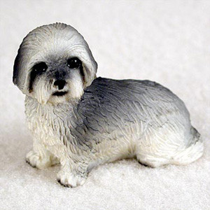 A detailed bonsai tree figurine of a Lhasa Apso dog with a gray and white "sport cut" coat. The figurine depicts the dog in a resting position, facing forward with a gentle expression, dark eyes, and a small black nose. The fur is meticulously sculpted to show texture, with salt-and-pepper gray shading on the ears and back, transitioning into a bright white on the face, chest, and paws. This miniature ornament is designed to be placed at the base of a bonsai tree, adding a lifelike and whimsical element to a miniature indoor garden or display.