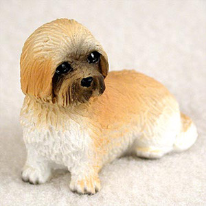 A detailed miniature figurine of a Lhasa Apso dog, specifically designed as a decorative accessory for a bonsai tree or indoor garden. The small dog figurine is depicted in a "sport cut" style, featuring textured fur that captures the breed's character. The coloring includes a warm tan or light brown shade across the top of its head and along its back, which fades into a clean white on its chest, belly, and four paws. Its expressive face is highlighted by dark brown and black markings around the muzzle and nose, surrounding two large, glossy black eyes. The figurine is shown in a seated pose, looking slightly to the left, and is photographed against a soft, neutral off-white background. This brown and white Lhasa Apso miniature serves as a charming addition to any bonsai tree display.