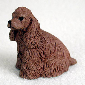 A detailed, chocolate brown American Cocker Spaniel figurine designed as a decorative accessory for a bonsai tree garden. The small miniature dog is shown in a seated pose, facing slightly to the left. Its entire body is covered in a sculpted texture that realistically represents the long, wavy coat of a spaniel, particularly on its iconic long, drooping ears and chest. The figurine features a short, blunt muzzle with a black nose and dark, glossy eyes that give it a gentle expression. It is set against a plain, light-colored background, highlighting the rich brown color and intricate detailing of the piece.