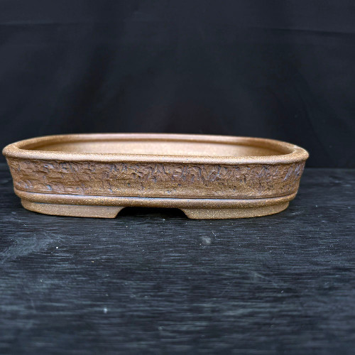 An 11-inch handmade ceramic bonsai pot by artist Jack Hoover, featuring a shallow oval design with a rich, earthy brown finish. The exterior of the pot is characterized by a deeply rugged, bark-like texture that mimics the appearance of weathered wood or stone, framed by a smooth, rounded top rim and a matching smooth horizontal band near the base. The clay body has a subtle, metallic sandy sheen that catches the light, adding depth to its artisanal appearance. Designed for professional bonsai display, the pot rests on a recessed foot with visible side cutouts for drainage and airflow, shown here on a dark wood-grain surface against a solid black background.
