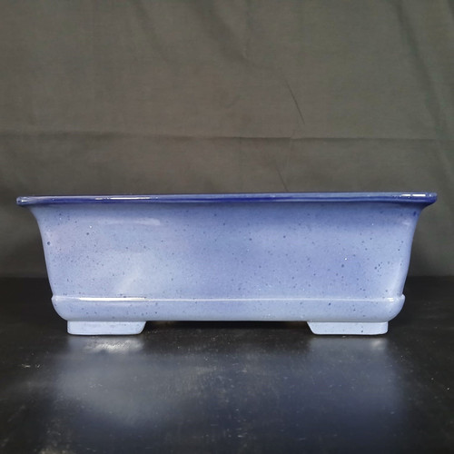 A side view of a 16-inch rectangular Yixing bonsai pot featuring a glazed periwinkle blue finish. The pot's surface is finely speckled with small, dark flecks, adding texture and depth to its soft lavender-blue hue. It has a pronounced top rim highlighted with a darker blue glaze that flares slightly outward. The pot is designed with a decorative ridge near the bottom and sits on four low, rectangular feet that provide a slight elevation. The ceramic vessel is photographed on a dark, smooth surface with subtle reflections, set against a dark, minimalist background that makes the light blue color pop.