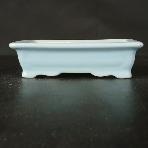 A side-view photograph of a 7-inch rectangular Yixing ceramic bonsai pot, model No. 2327C. The pot is finished in a serene, pale blue glaze with a soft, uniform sheen. Its design features a subtly flared rim and four short, elegantly curved feet that lift the base slightly off the surface. The pot is positioned on a dark, textured tabletop with a subtle reflection of the light blue glaze visible below. The background is a solid, dark, non-distracting color, which emphasizes the pot's clean rectangular lines and delicate color. This professional-grade Yixing clay pot is designed for aesthetic beauty and functional drainage, making it an ideal vessel for various small bonsai tree species.