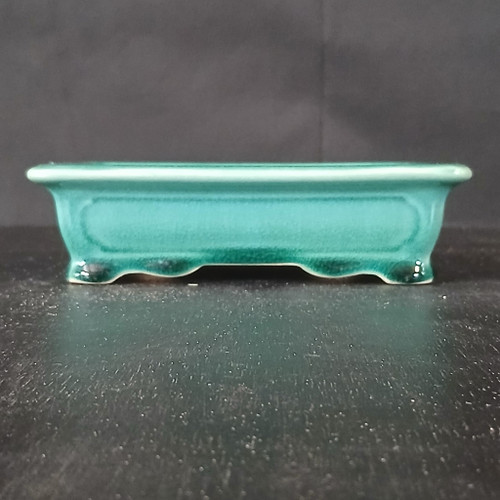 A side-view, eye-level photograph of a 7-inch glazed rectangle Yixing ceramic bonsai pot. The pot is finished in a vibrant, glossy teal green glaze that shows a subtle crackle texture and deeper green pooling near the base and corners. It has a classic rectangular shape with rounded corners and a slightly flared, thick upper rim. The front face features a shallow, recessed rectangular panel, adding depth to the design. Supporting the pot are four small, integrated feet that are part of a decorative, wavy scalloped bottom edge. The ceramic pot is centered on a dark, textured surface against a soft-focus, dark grey background, making the bright teal color and elegant craftsmanship stand out.