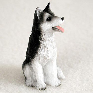 A detailed miniature Siberian Husky figurine designed as a decorative accessory for a bonsai tree. The small figurine is shown sitting down on a neutral, light-colored surface, looking off to the right with an alert and friendly expression. 

The husky has a classic black and white coat with sculpted fur texture. Its back, the top of its head, and its pointed ears are black, while its chest, underbelly, legs, and face are bright white. The dog’s mouth is slightly open, revealing a small pink tongue as if it is panting or smiling, and it features dark, attentive brown eyes. This miniature Siberian Husky figurine is crafted to add a touch of personality and life to a bonsai landscape or miniature garden.